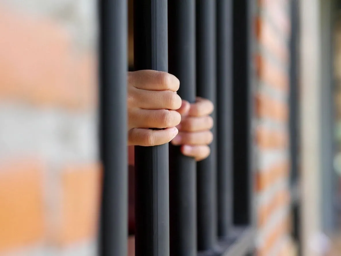 A hand grips a vertical bar of a jail cell window.