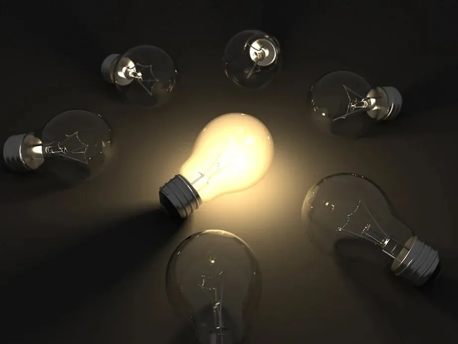 One lit incandescent light bulb among several unlit bulbs on a dark surface