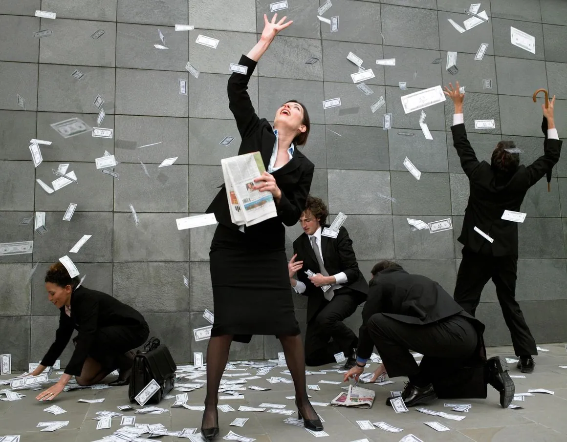 Business people in dark suits reaching up and crouching to gather banknotes falling around them