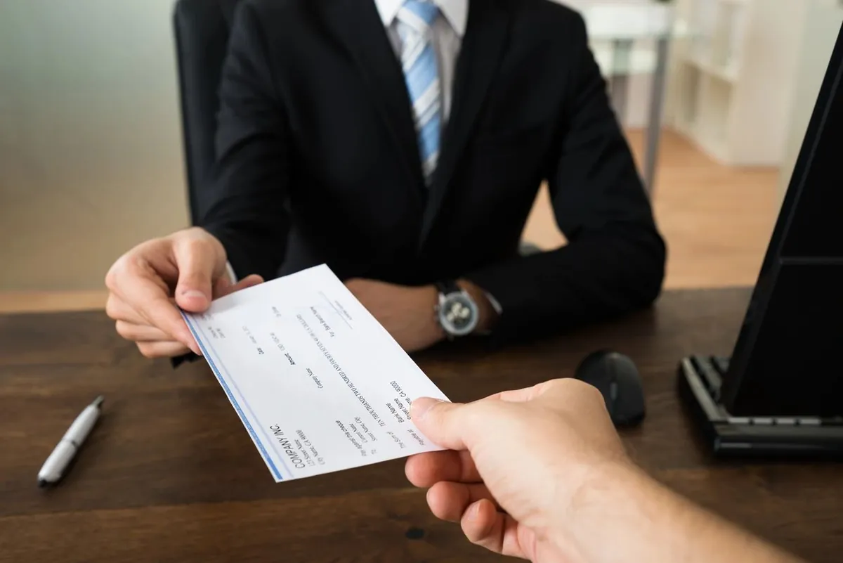 A man in a suit hands a paper check across a desk to another person
