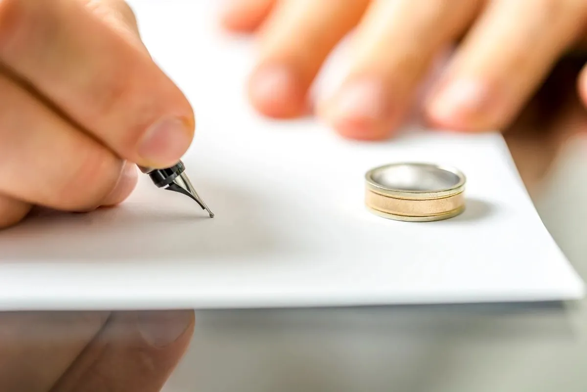 A hand signing a paper with a fountain pen, with a wedding ring beside the paper.