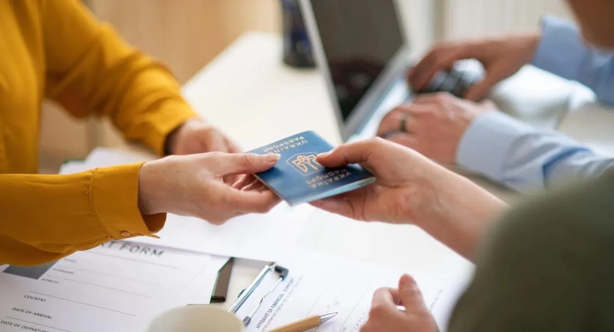 A person handing a card to another person across a desk, with a form and a laptop on the desk.