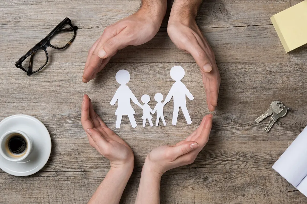 Two pairs of hands cupped around a paper-cut family on a wooden desk with glasses, a coffee cup and keys.