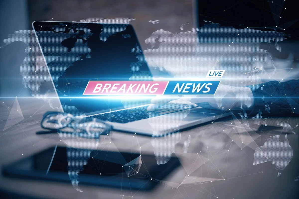 A laptop on a desk showing a breaking-news banner over a world map, with glasses and a tablet beside it