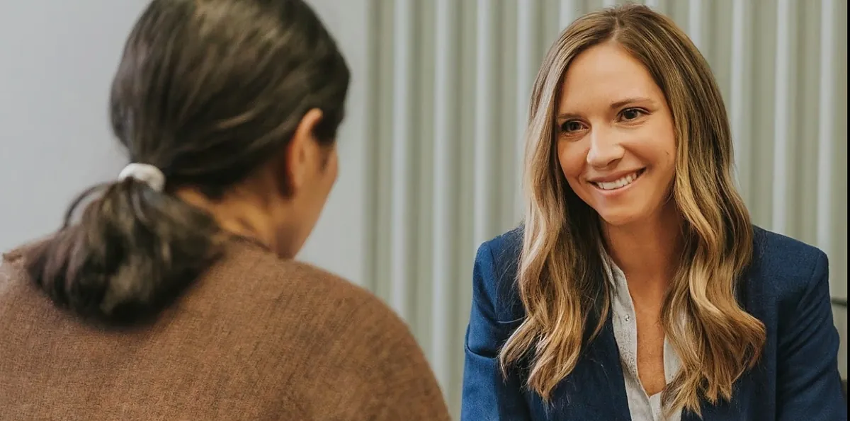Two women sitting across a table and talking; one smiles and wears a blue blazer, the other is seen from behind.