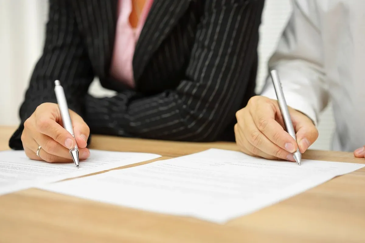 Two people at a table, each holding a pen over a printed agreement.