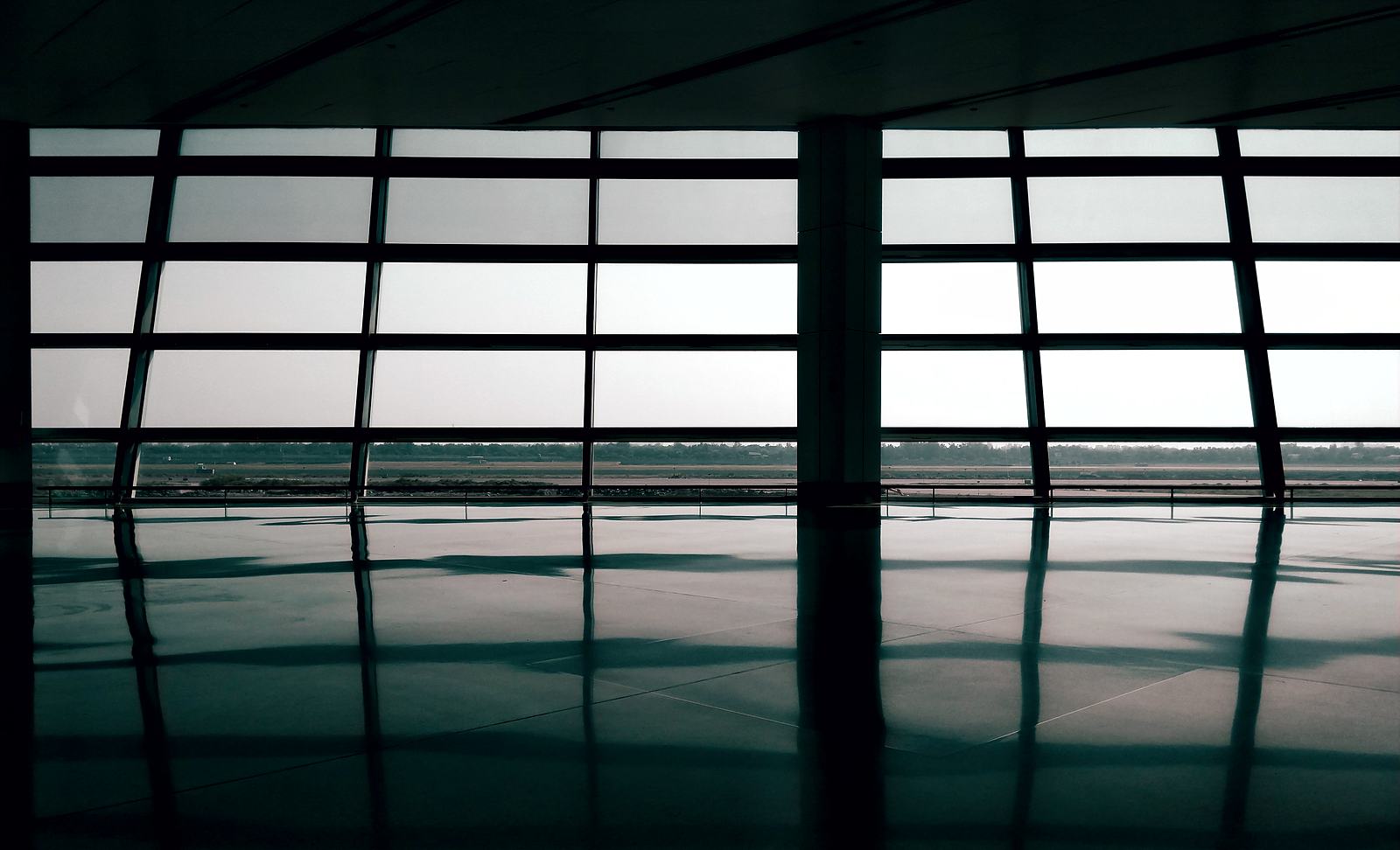 A dark airport terminal hall with a wall of floor-to-ceiling windows, whose grid is mirrored in the polished floor.