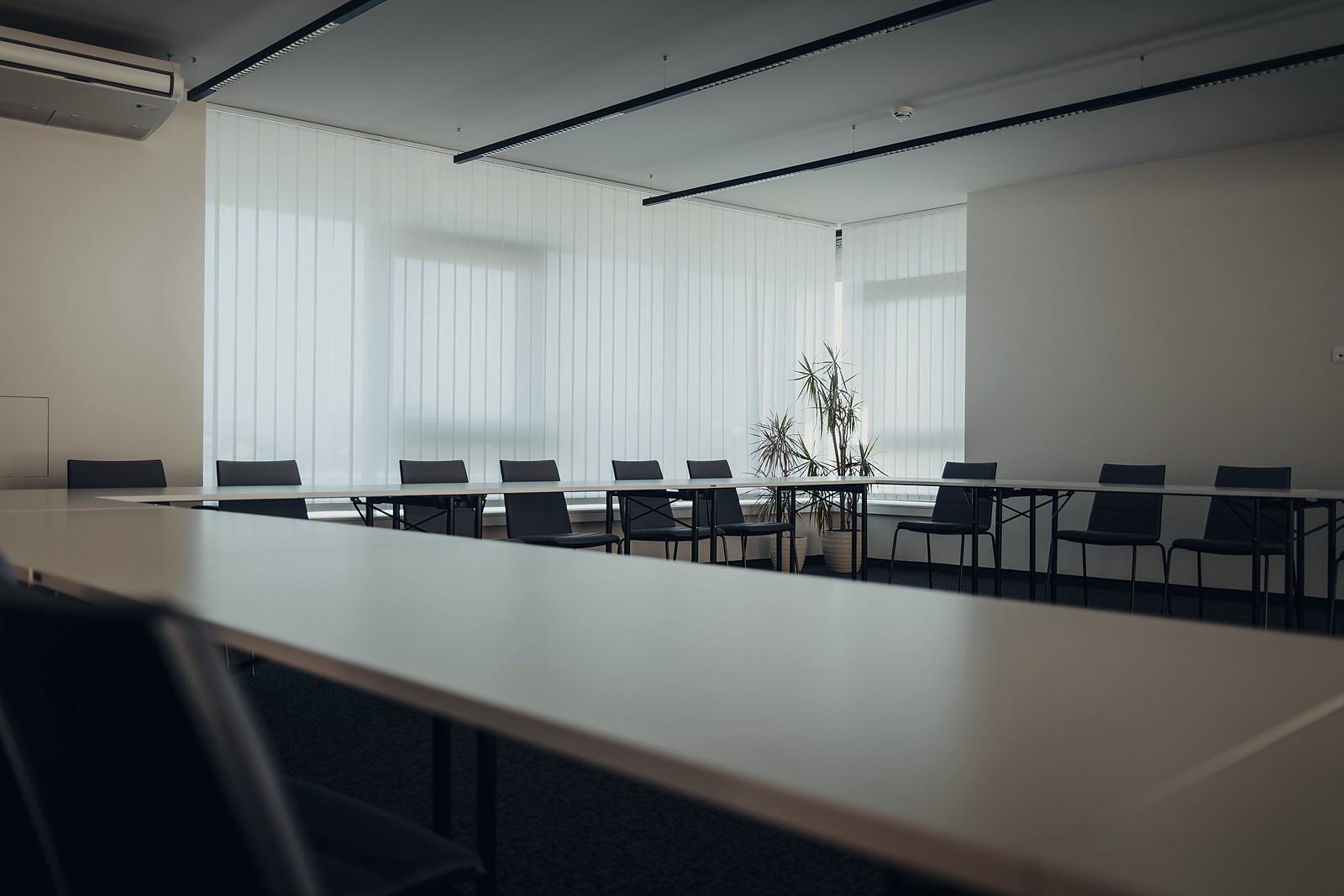 An empty meeting room with a long table, dark chairs and a window wall behind white vertical blinds.