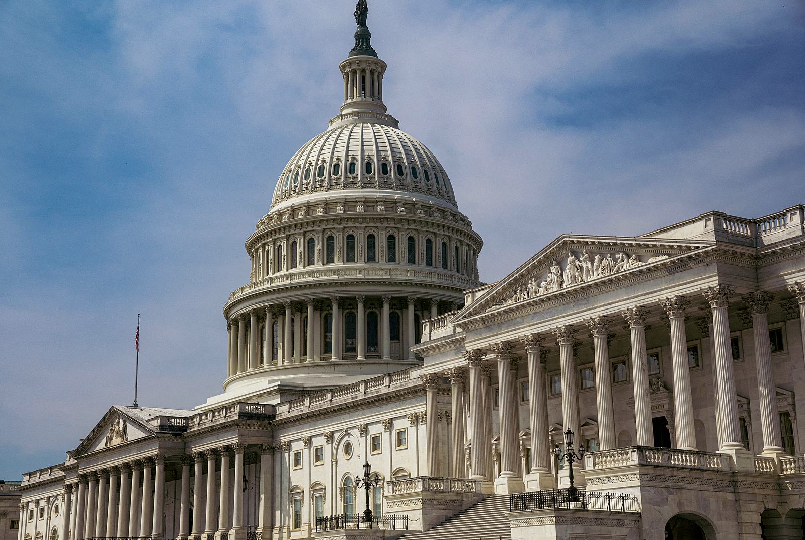 The dome and a colonnaded wing of the U.S. Capitol in Washington, D.C., under a blue sky with thin cloud.