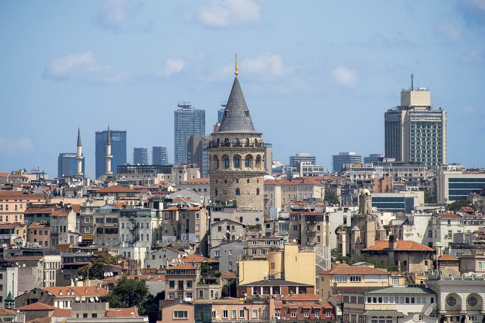 Istanbul's Galata Tower rising above the old rooftops of the Galata hillside, with modern office towers behind it.