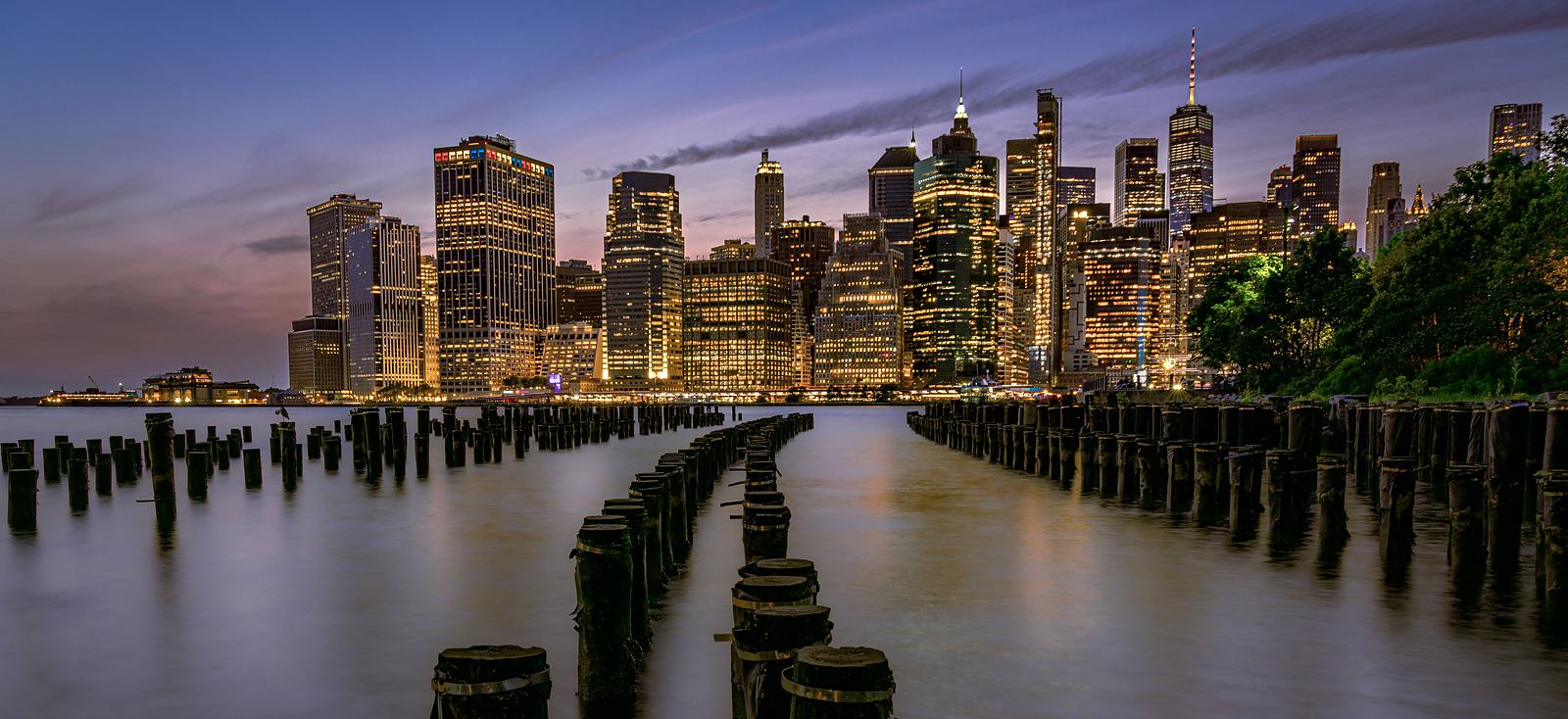 Lower Manhattan at dusk seen from Brooklyn, its office towers lit above calm water and rows of old pier pilings.